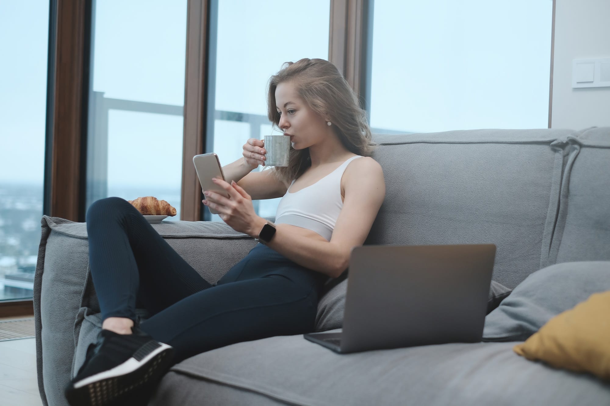 Young woman drinks coffee sitting on sofa with laptop and telephone